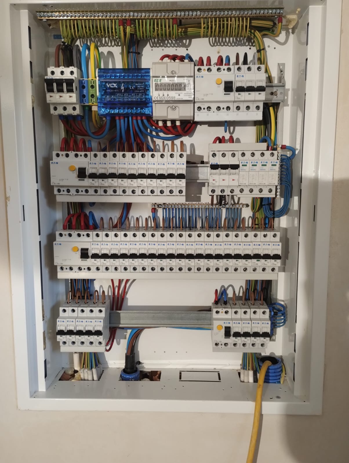 Rozdzielnica elektryczna w białej obudowie, widoczne liczne zabezpieczenia, przewody w różnych kolorach i moduł VOX. Całość schludnie ułożona.