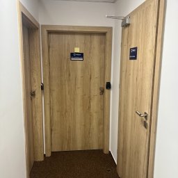 Mivol - Korytarz biurowy z trzema drewnopodobnymi drzwiami, z czego jedne wyposażone w samozamykacz i oznaczone szyldem 'Policja'.