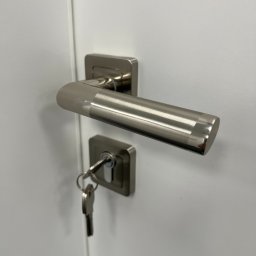 Mivol - Szczegółowe ujęcie srebrnej klamki i zamka z kluczem w białych drzwiach wewnętrznych.