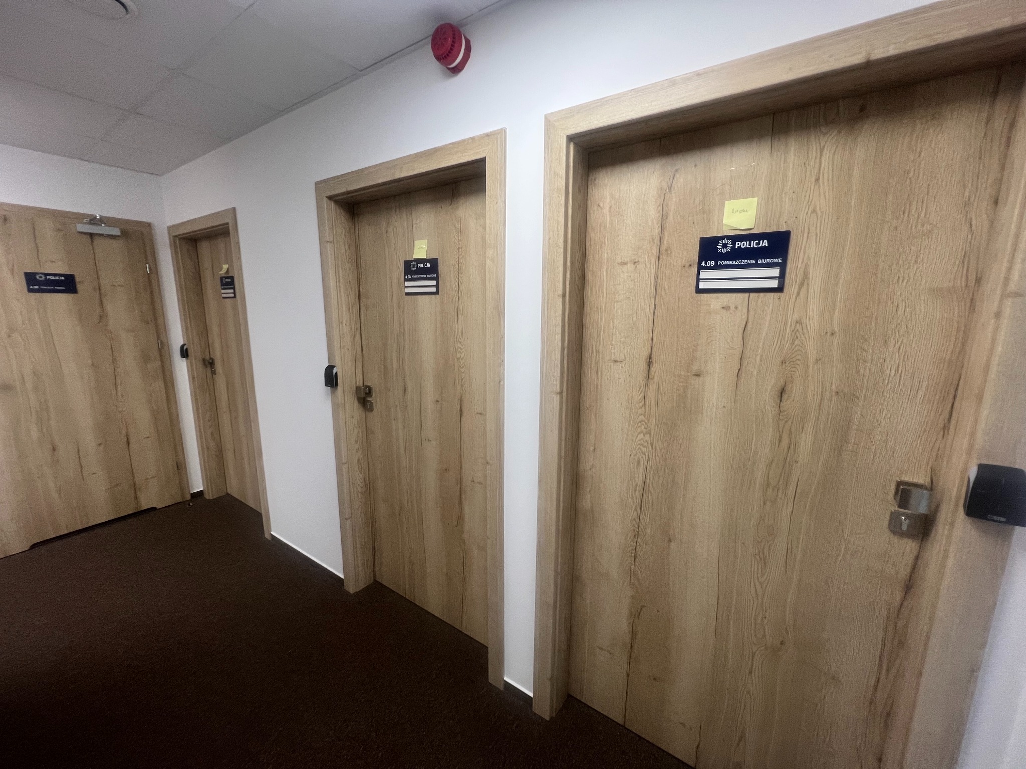 Korytarz z trzema drewnianymi drzwiami z tabliczkami informacyjnymi 'Policja' i numerami pomieszczeń, widoczne brązowe wykładziny podłogowe.