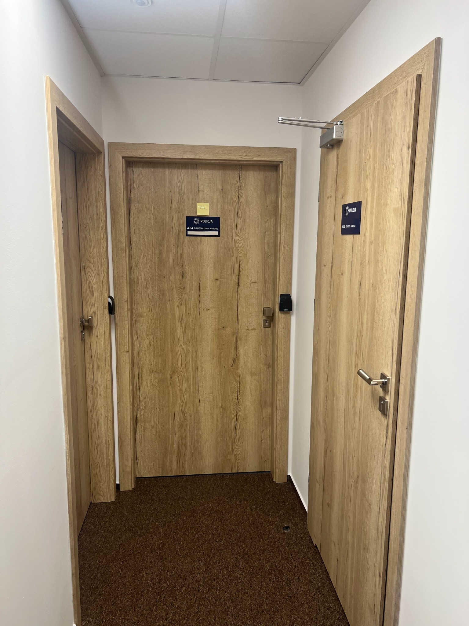 Korytarz biurowy z trzema drewnopodobnymi drzwiami, z czego jedne wyposażone w samozamykacz i oznaczone szyldem 'Policja'.