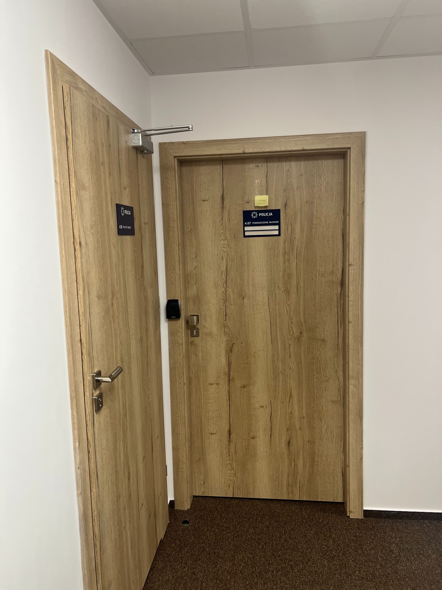 Dwa drewnopodobne drzwi wewnętrzne w biurze policji, jedne z samozamykaczem i kartą dostępu, drugie uchylone z tabliczką informacyjną.