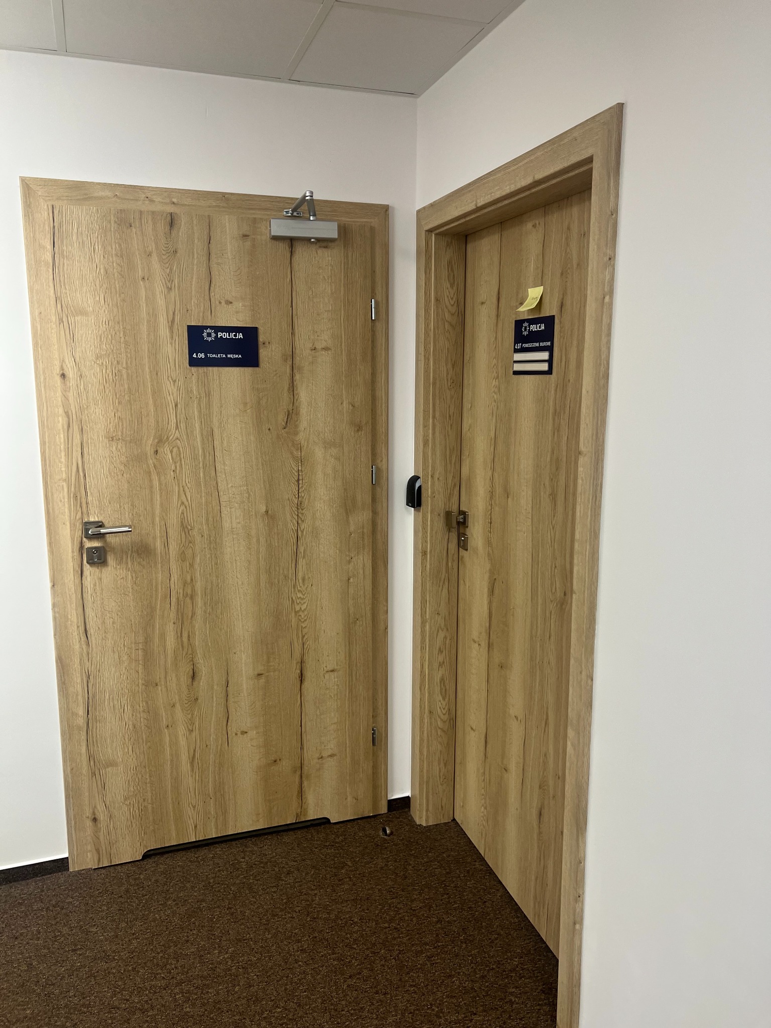 Dwie drewniane drzwi wewnętrzne w biurze policji w Warszawie, z tabliczkami informacyjnymi, widoczne elementy montażowe i brązowa wykładzina podłogowa.