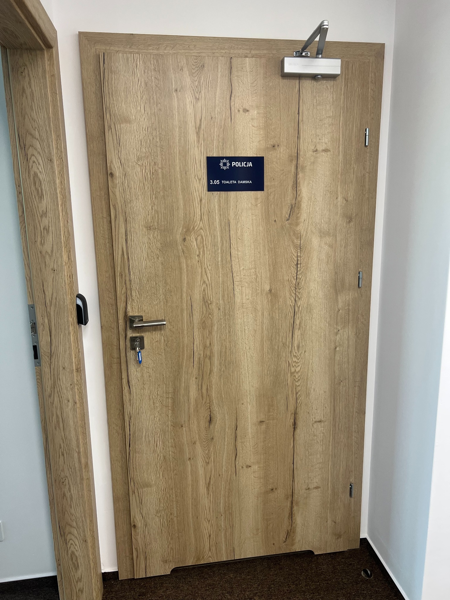 Beżowa płycinowa okleina na drzwiach z zamontowanym samozamykaczem i tabliczką 'Policja Toaleta Damska 3.05', widoczne klucze w zamku.