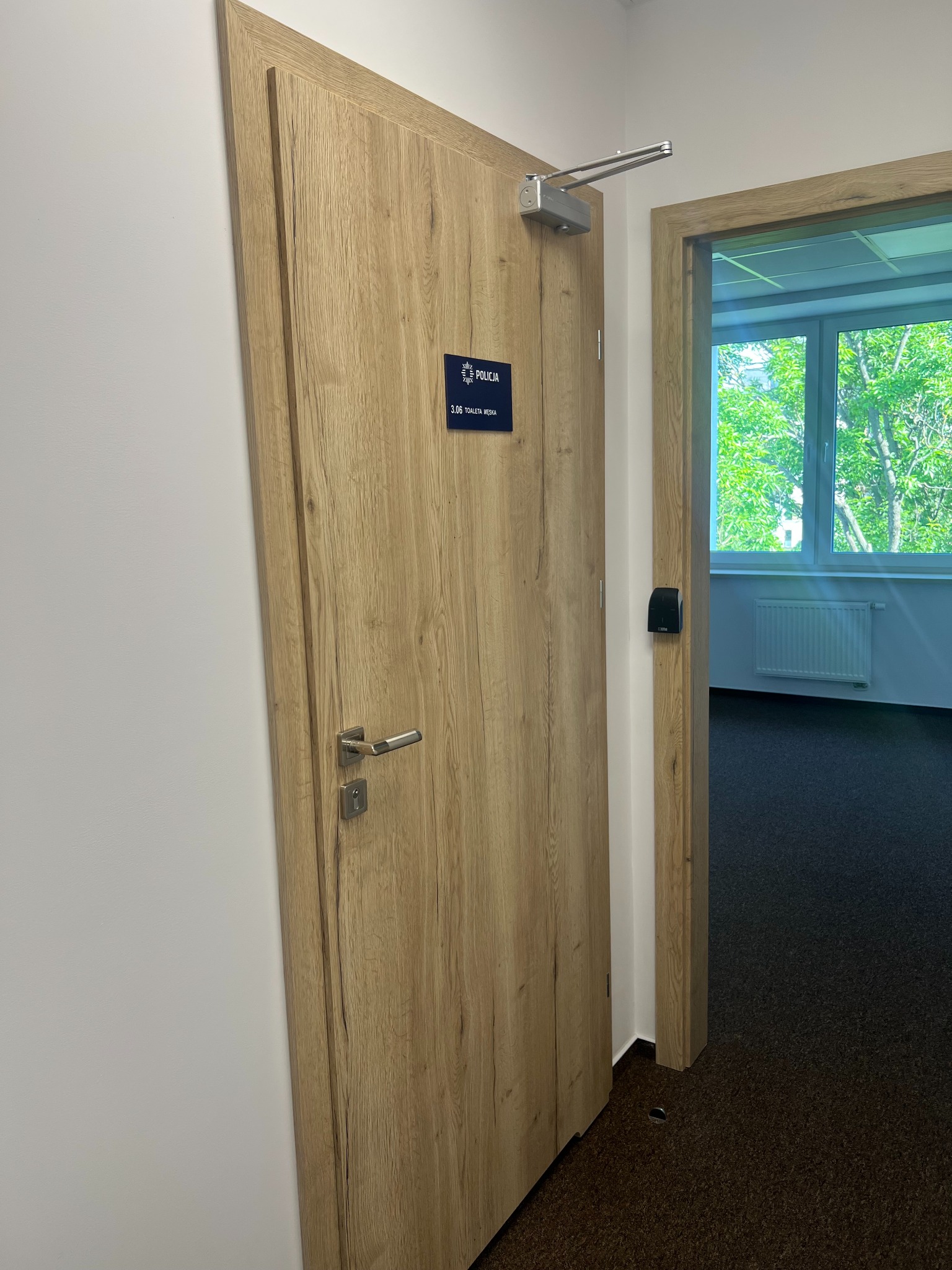 Dębowa okleina drzwi z samozamykaczem i tabliczką 'Policja 3.06 Toaleta Męska', widok na biuro z oknem i grzejnikiem