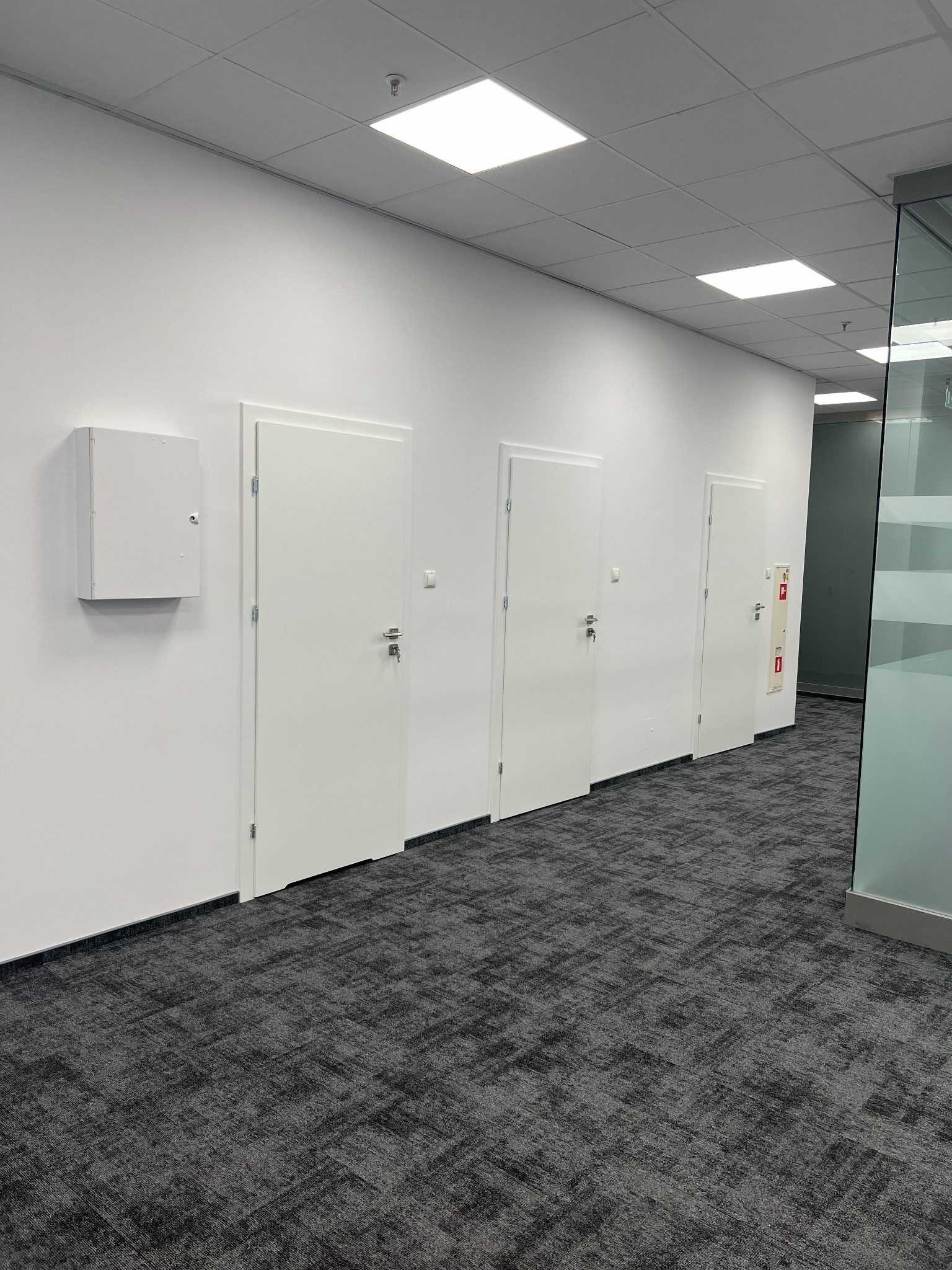 Korytarz biurowy z trzema białymi drzwiami i szarą wykładziną dywanową, widoczna szafka elektryczna i gaśnica na ścianie.