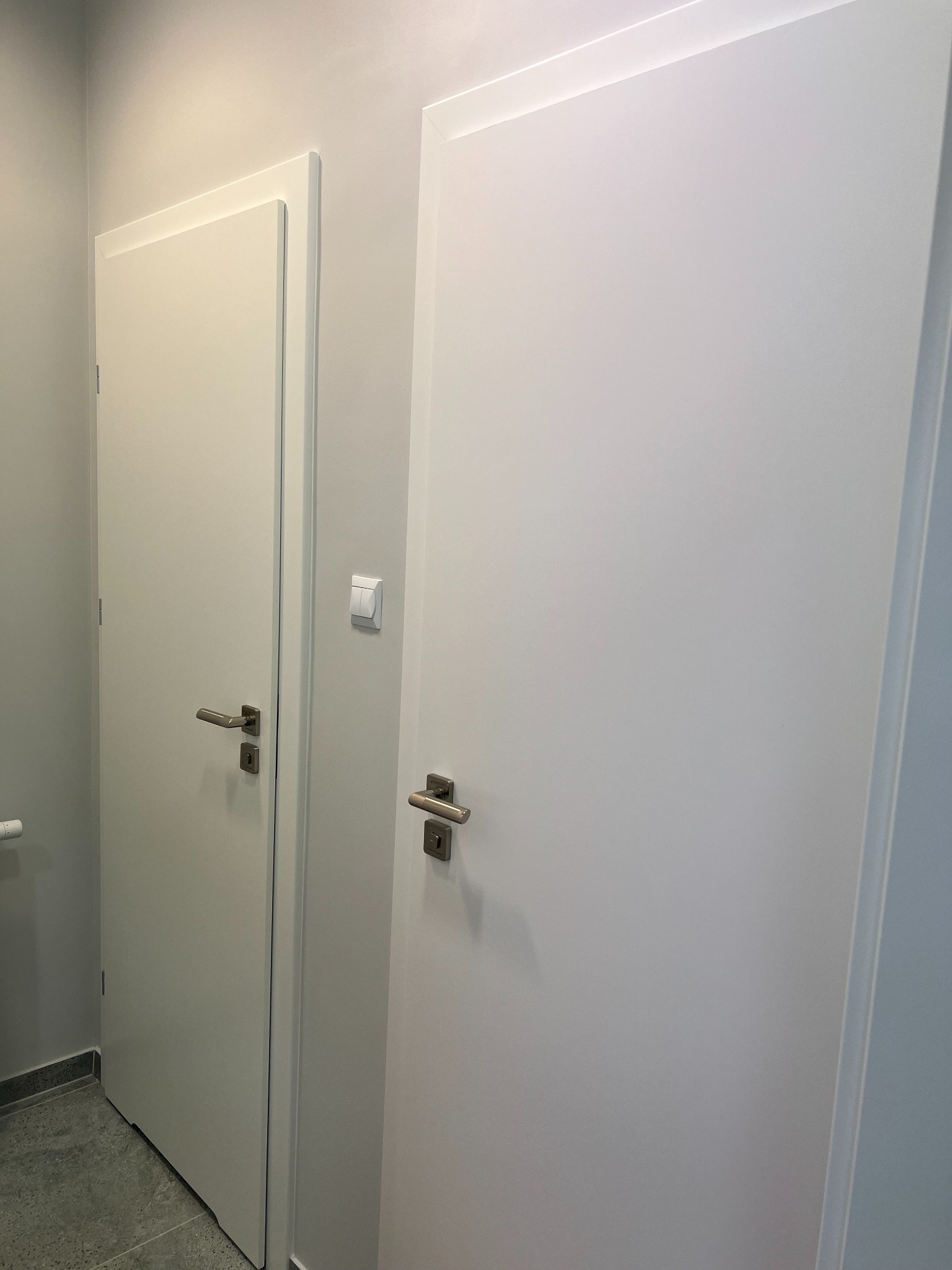 Dwa białe, minimalistyczne drzwi wewnętrzne z metalowymi klamkami, jedno z widocznym włącznikiem światła na ścianie, drugie z fragmentem grzejnika w rogu, na tle szarej ściany i szarej podłogi.