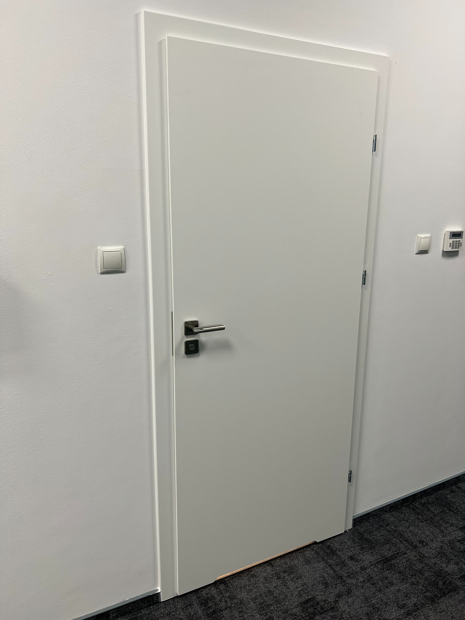 Zamknięte, białe skrzydło drzwiowe z ościeżnicą w minimalistycznym stylu, widoczne zawiasy i klamka na tle białej ściany z włącznikiem i panelem alarmowym.