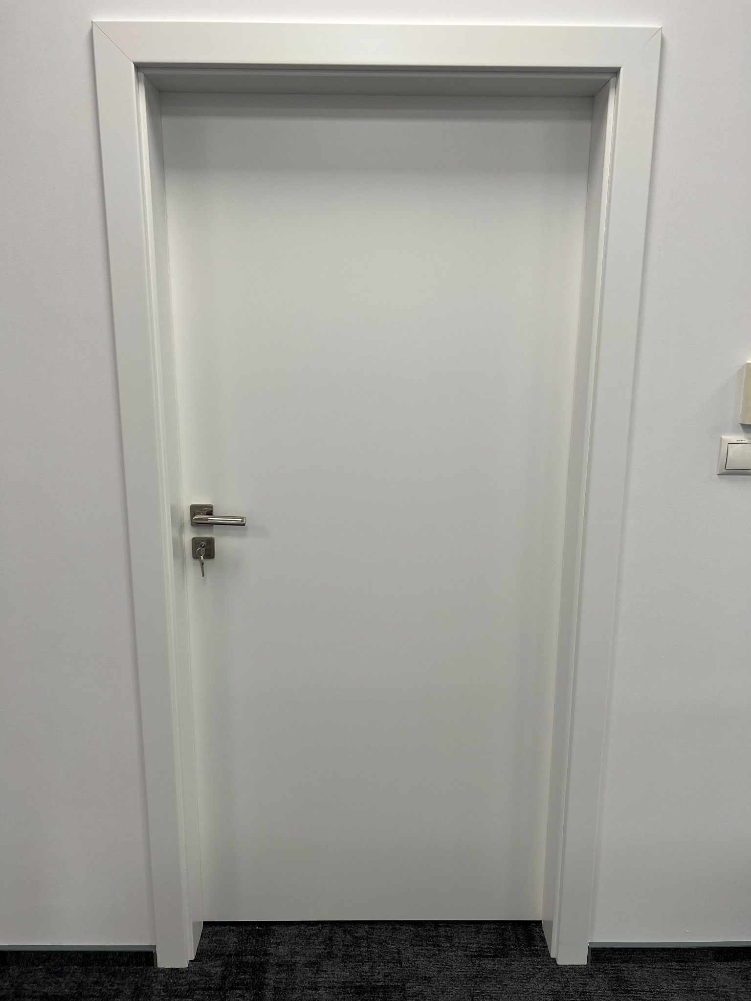 Zamknięte, białe skrzydło drzwiowe z prostą ościeżnicą i metalową klamką z kluczem w zamku, widoczne na tle białej ściany i szarej wykładziny podłogowej.