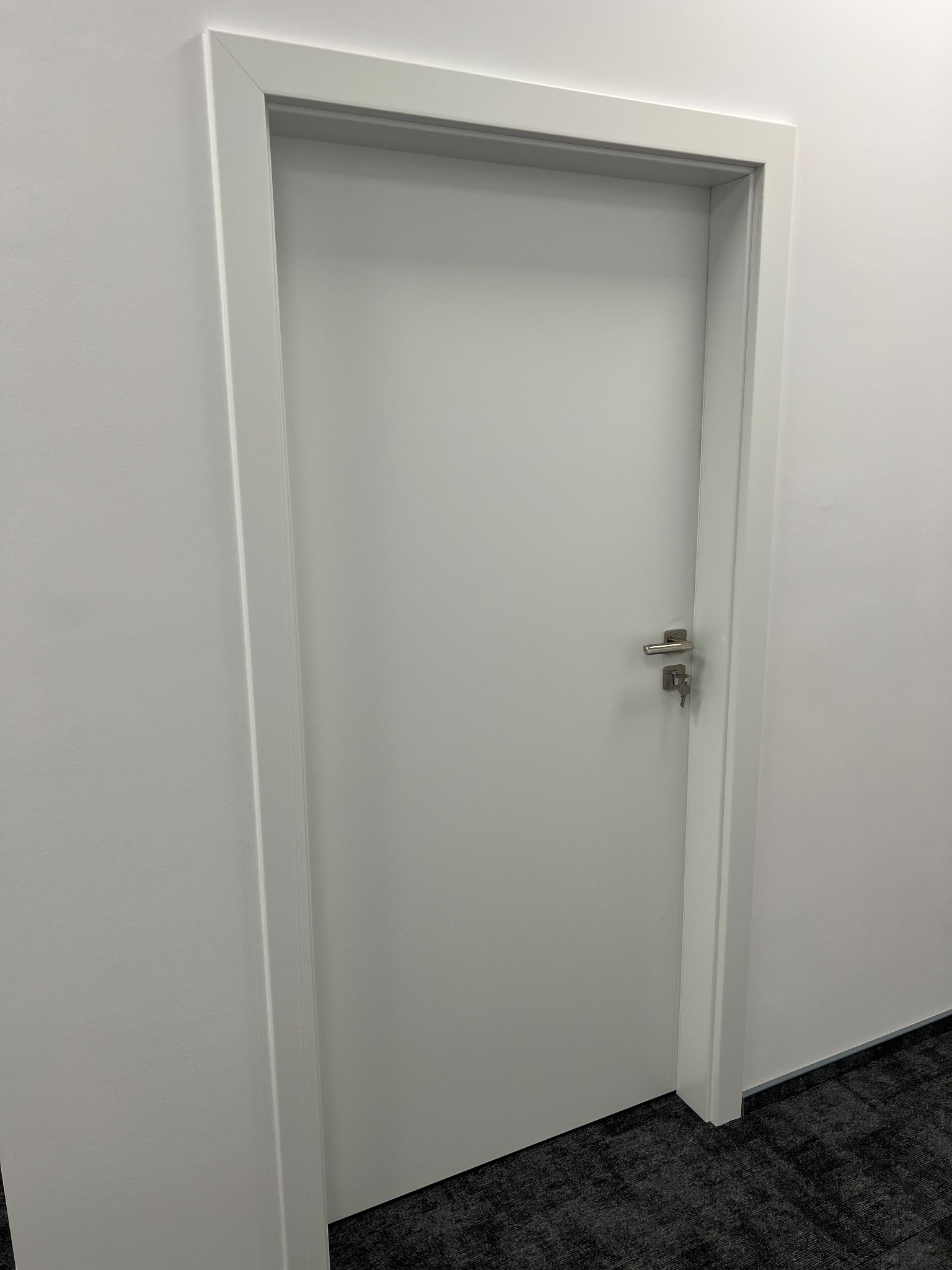 Zamknięte, białe drzwi z ościeżnicą w białym kolorze, z kluczem w zamku, na tle białej ściany i szarej wykładziny.