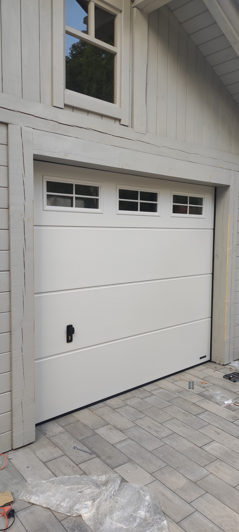 Biała brama garażowa segmentowa z trzema małymi okienkami w górnej sekcji, zamontowana w jasnoszarym budynku z drewnianą elewacją, widoczne narzędzia i folia ochronna na kostce brukowej.