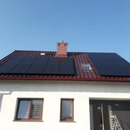 Częściowy widok domu jednorodzinnego z brązową dachówką i panelami fotowoltaicznymi zamontowanymi na dachu, widoczny ceglany komin i okno dachowe, na dole ściana budynku z oknami i częściowo...