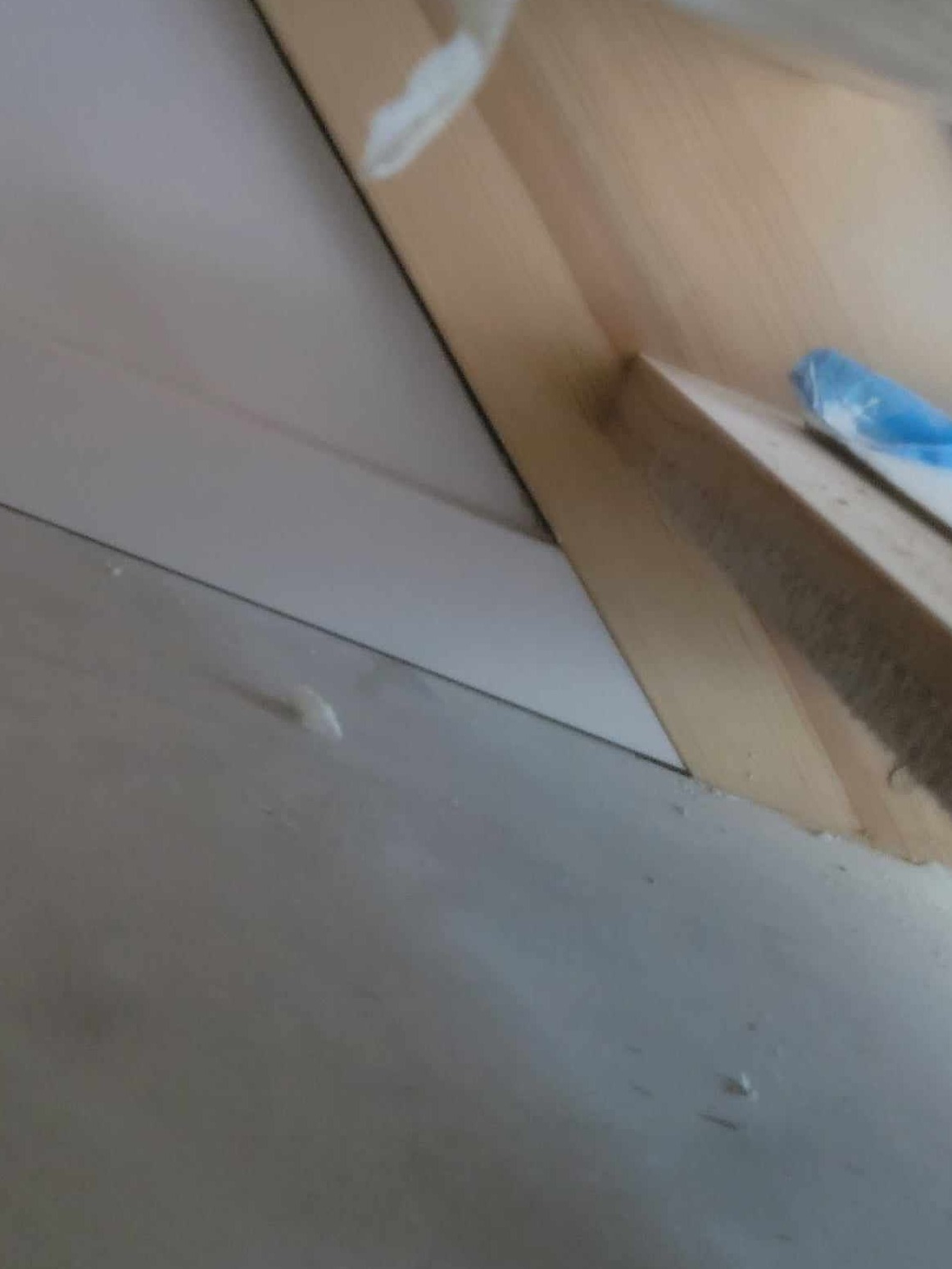 Fragment podłogi z ułożonymi panelami, widoczne drobne zabrudzenia i listwa przypodłogowa w trakcie montażu, z niebieską folią ochronną.