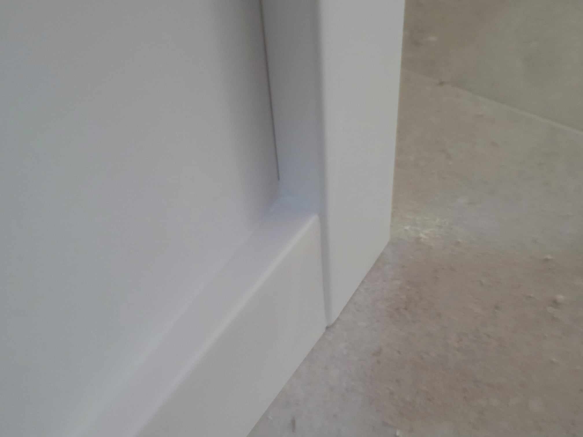 Detal narożnika ściany z białą listwą przypodłogową i fragmentem jasnej, matowej podłogi.