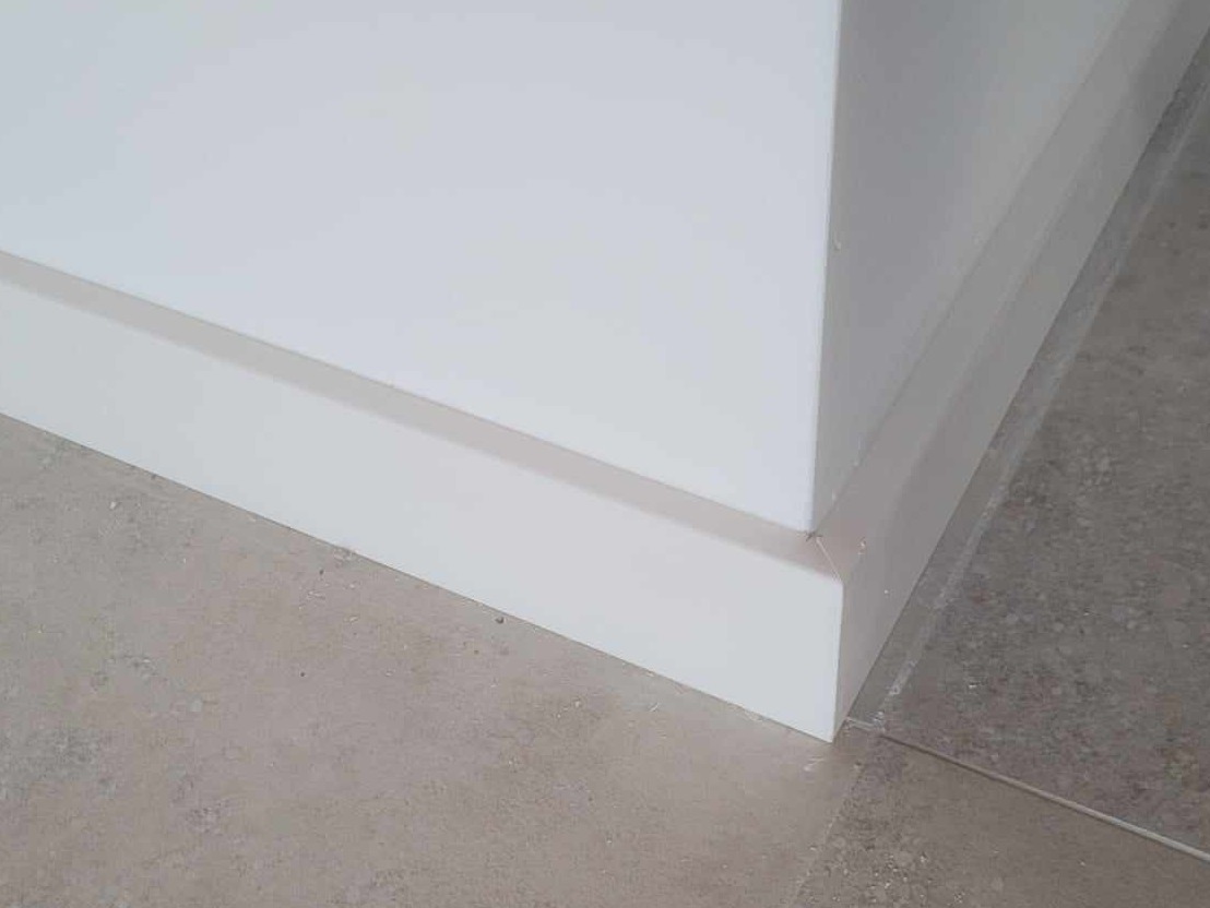 Detal narożnika pomieszczenia z ułożonymi, białymi listwami przypodłogowymi na szarych płytkach podłogowych.