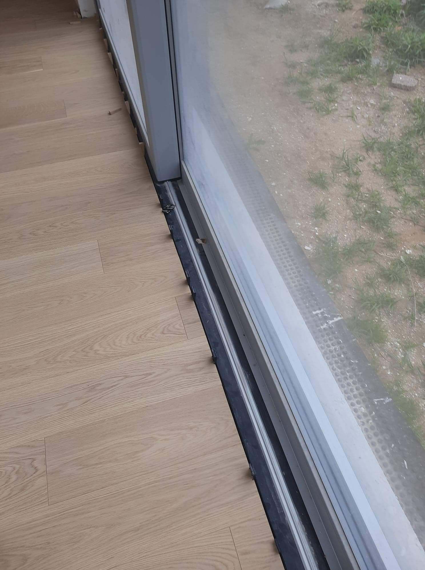 Fragment podłogi z jasnych paneli, widoczne kliny dystansowe przy szarej ramie okna balkonowego; na zewnątrz widoczny grunt z rzadką roślinnością.