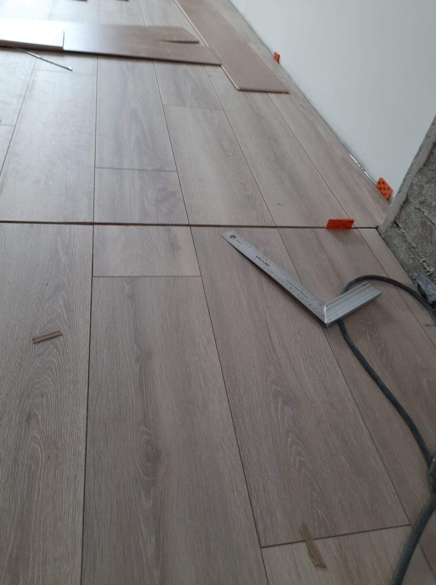Fragment podłogi z ułożonymi panelami laminowanymi w odcieniach szarości, widoczny kątownik i elementy dystansowe przy ścianie, praca w trakcie.