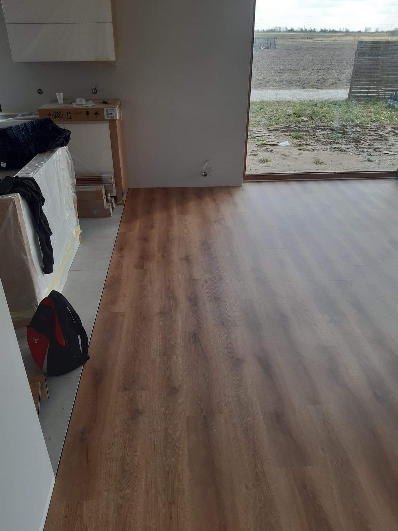 Nowo ułożone panele podłogowe o ciepłym, brązowym odcieniu, z widocznymi paczkami paneli w tle, częściowo zakrytymi folią, oraz plecakiem leżącym na podłodze obok