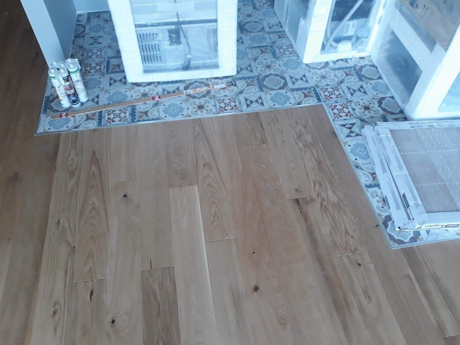 Fragment podłogi z ułożonych paneli drewnianych, widoczne puszki z pianką montażową przy ścianie z dekoracyjnymi płytkami i zabezpieczone folią meble.