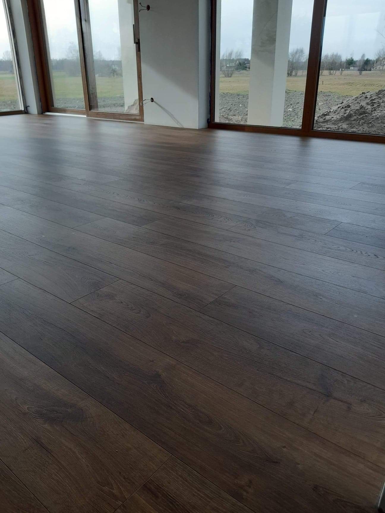 Ciemne panele podłogowe ułożone w nowym, jasnym pomieszczeniu z dużymi oknami, widok na zewnątrz na pole i hałdę ziemi.