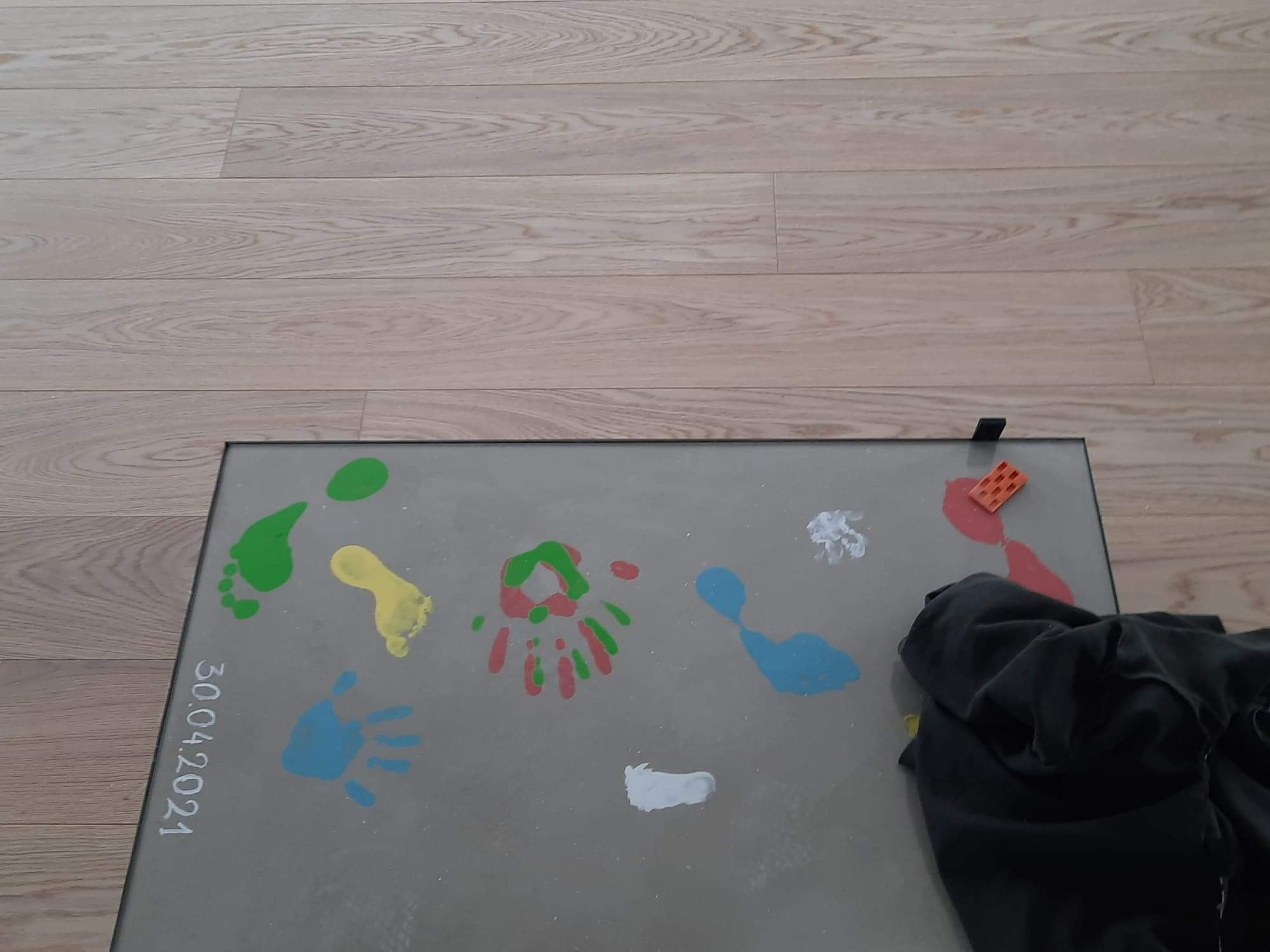 Świeżo położone panele podłogowe z dziecięcymi odciskami stóp i dłoni pomalowanymi farbami w różnych kolorach, data 30.04.2021 zapisana na krawędzi.