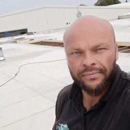 MajsterHaus SŁAWOMIR PLACEK - Mężczyzna na dachu budynku przemysłowego w trakcie prac dekarskich, widoczne narzędzia i elementy konstrukcji dachu. Białe pokrycie dachowe.