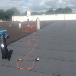 MajsterHaus SŁAWOMIR PLACEK - Prace dekarskie na dachu płaskim: papa termozgrzewalna, butla gazowa, wózek z rolką papy. Część dachu pokryta nową warstwą, druga w starszym kolorze. Słoneczny dzień.