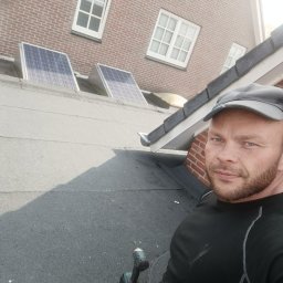 MajsterHaus SŁAWOMIR PLACEK - Mężczyzna w czarnej koszulce na dachu z papy, w tle dwa panele słoneczne, okna i fragment ceglanej ściany. W ręku wkrętarka.