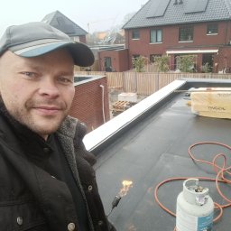 MajsterHaus SŁAWOMIR PLACEK - Dekarz w trakcie pracy na dachu płaskim, używa palnika do zgrzewania papy. W tle widoczne budynki mieszkalne i fragment krajobrazu miejskiego.