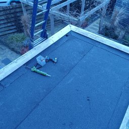 MajsterHaus SŁAWOMIR PLACEK - Płaski dach pokryty papą z widocznymi narzędziami (wiertarka, pojemnik na wkręty, tuba z uszczelniaczem) i drabiną w tle. Widoczne pęknięcia papy.