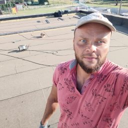 MajsterHaus SŁAWOMIR PLACEK - Pracownik na dachu z papy termozgrzewalnej w trakcie prac. Widoczne narzędzia i materiały. W tle zabudowa wiejska i plac zabaw.