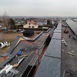 MajsterHaus SŁAWOMIR PLACEK - Widok z góry na dach w trakcie wymiany, z widocznymi elementami konstrukcyjnymi, materiałami budowlanymi i fragmentem zabudowy miejskiej w tle.