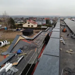 Widok z góry na dach w trakcie wymiany, z widocznymi elementami konstrukcyjnymi, materiałami budowlanymi i fragmentem zabudowy miejskiej w tle.