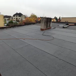 Nowe, płaskie pokrycie dachu z papy termozgrzewalnej z kominem. W tle zabudowa jednorodzinna i pochmurne niebo. Widoczny zwój papy.