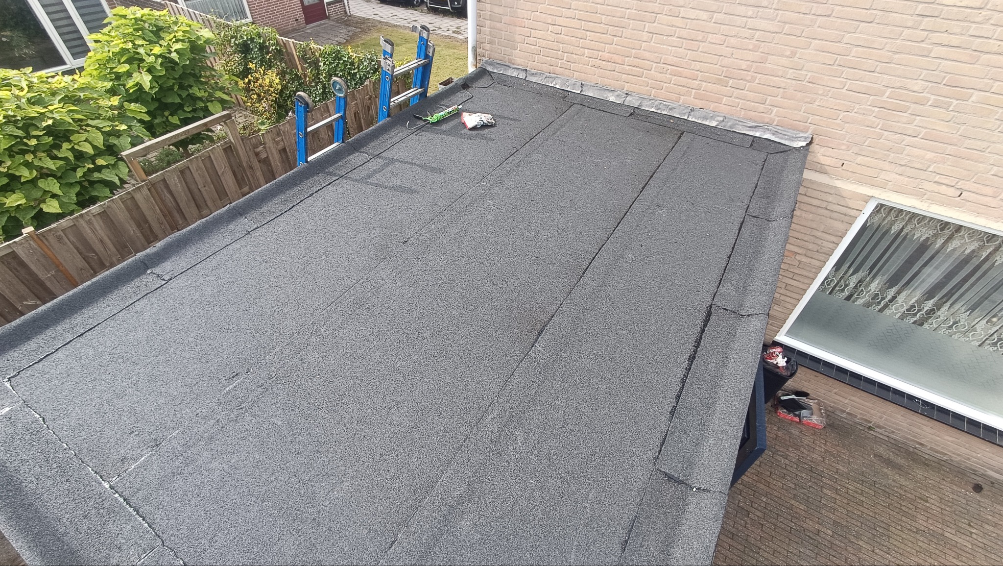 Szczegółowe ujęcie płaskiego dachu pokrytego papą, z drabiną opartą o ogrodzenie i narzędziami dekarskimi na powierzchni. Widok z góry.