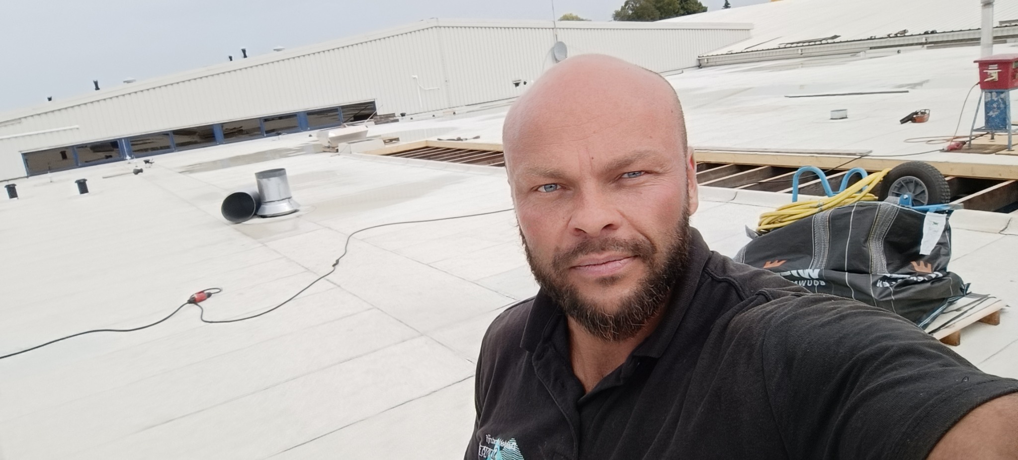 Mężczyzna na dachu budynku przemysłowego w trakcie prac dekarskich, widoczne narzędzia i elementy konstrukcji dachu. Białe pokrycie dachowe.