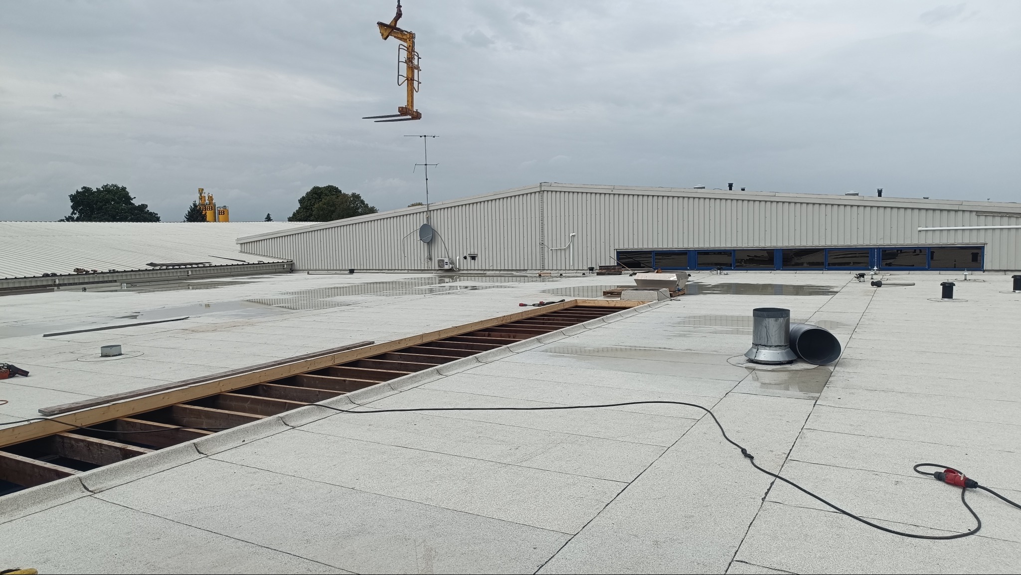 Płaski dach budynku przemysłowego w trakcie naprawy, z widoczną konstrukcją drewnianą i dźwigiem w tle, w pochmurny dzień. Widoczne narzędzia i elementy wentylacyjne.