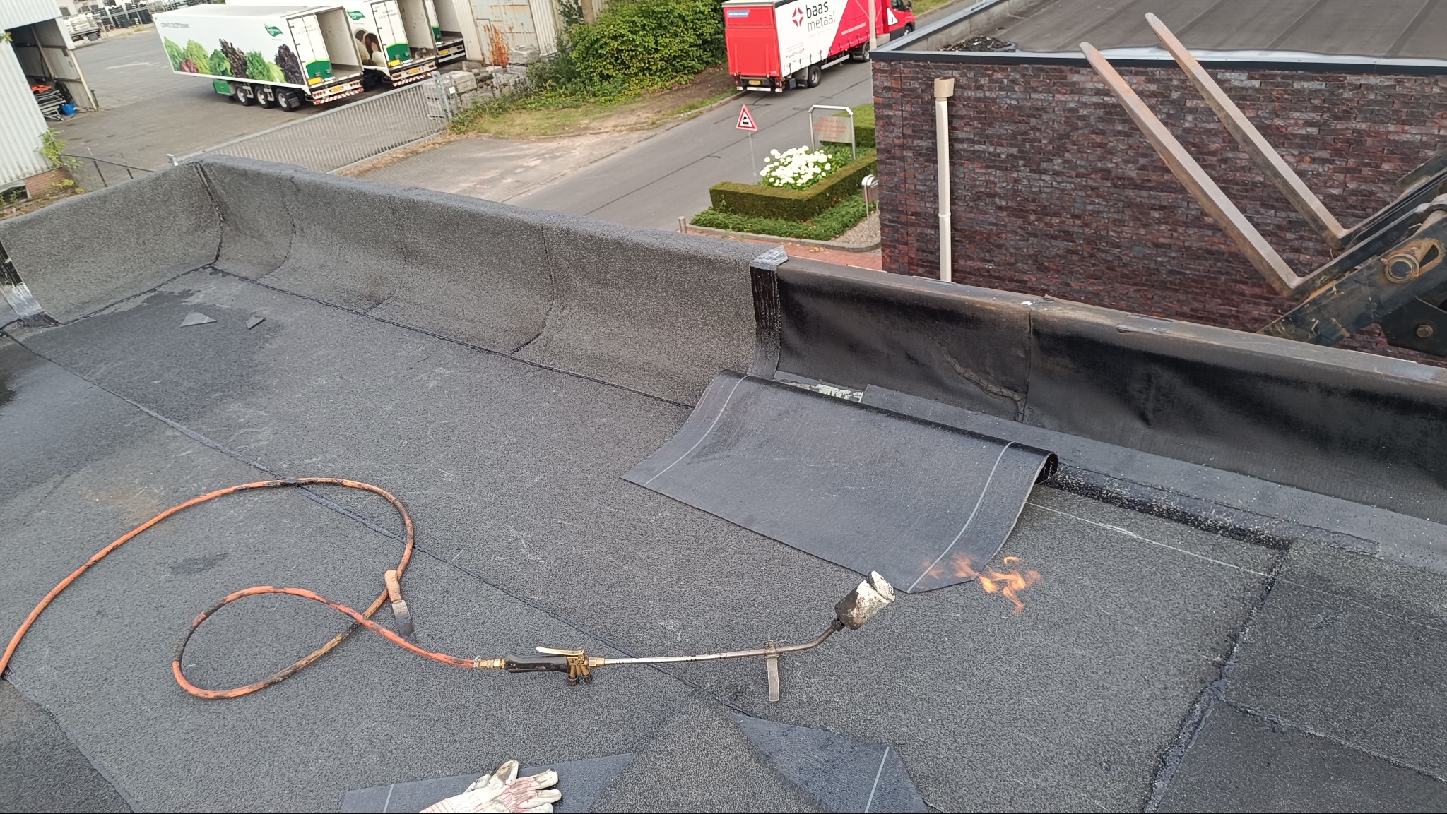 Praca na dachu płaskim: palnik dekarski w użyciu podczas zgrzewania papy, widoczne narzędzia i fragment dachu. W tle budynki i pojazdy.