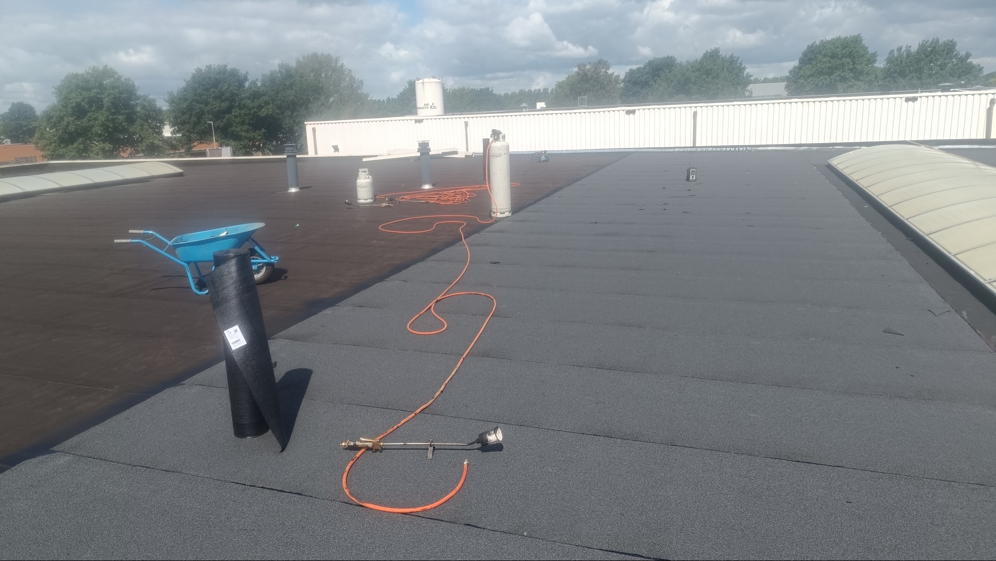 Prace dekarskie na dachu płaskim: papa termozgrzewalna, butla gazowa, wózek z rolką papy. Część dachu pokryta nową warstwą, druga w starszym kolorze. Słoneczny dzień.