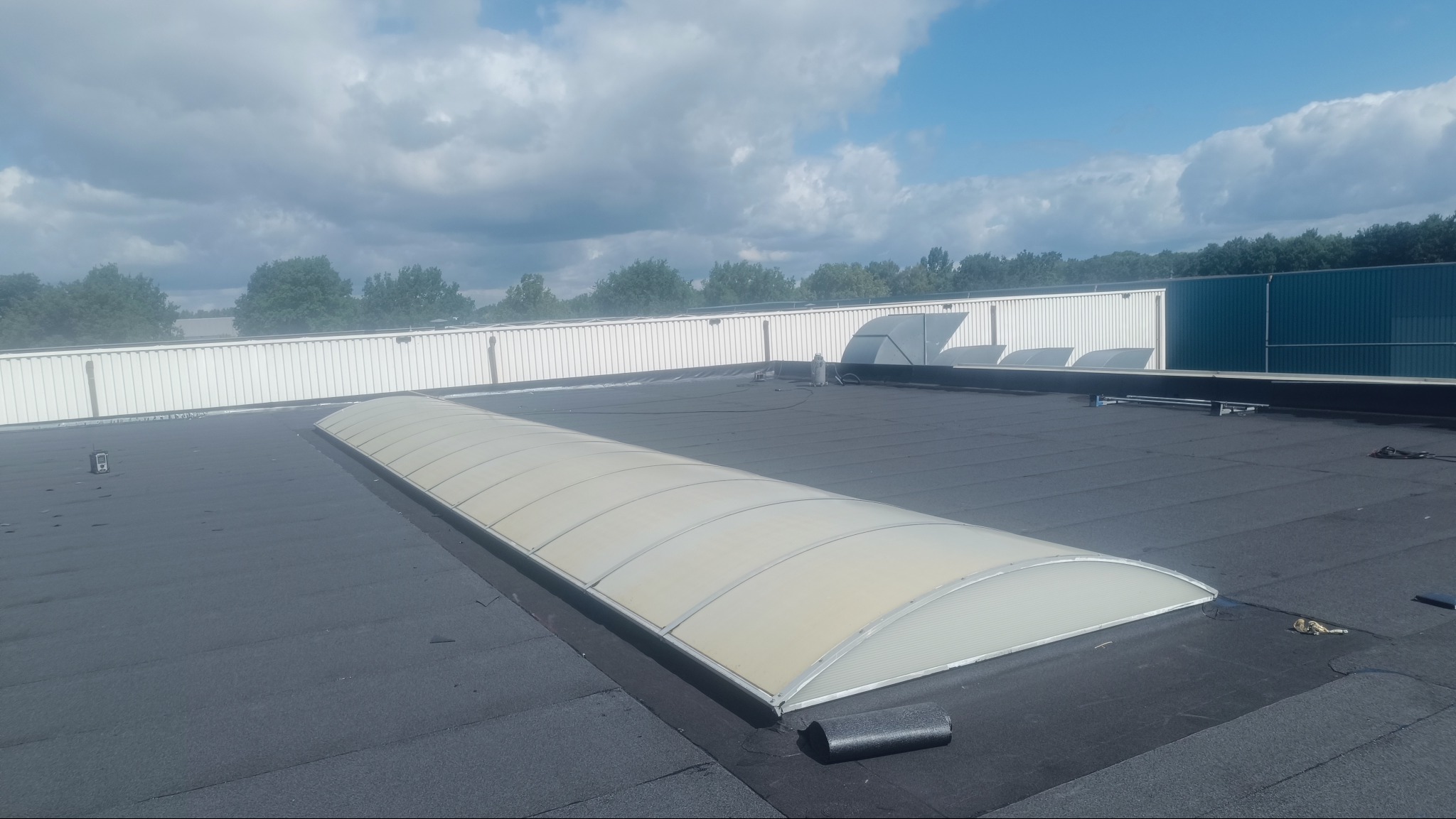 Dach pokryty czarną papą termozgrzewalną z zamontowanym świetlikiem tunelowym. W tle biała ściana hali i błękitne niebo z chmurami.