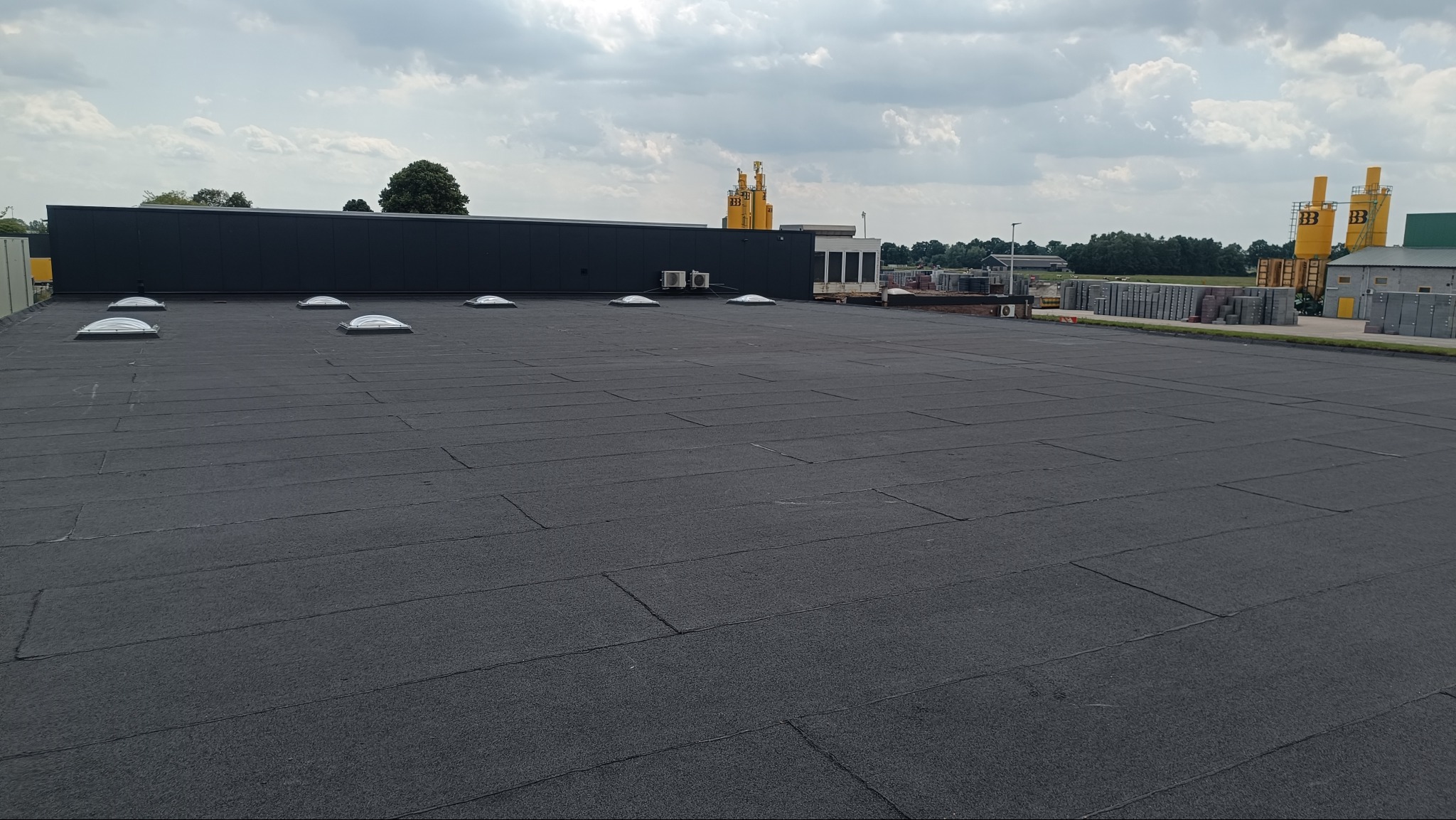 Wykończony, płaski dach z papy termozgrzewalnej na budynku przemysłowym z świetlikami. W tle widoczne silosy i składowane materiały budowlane.