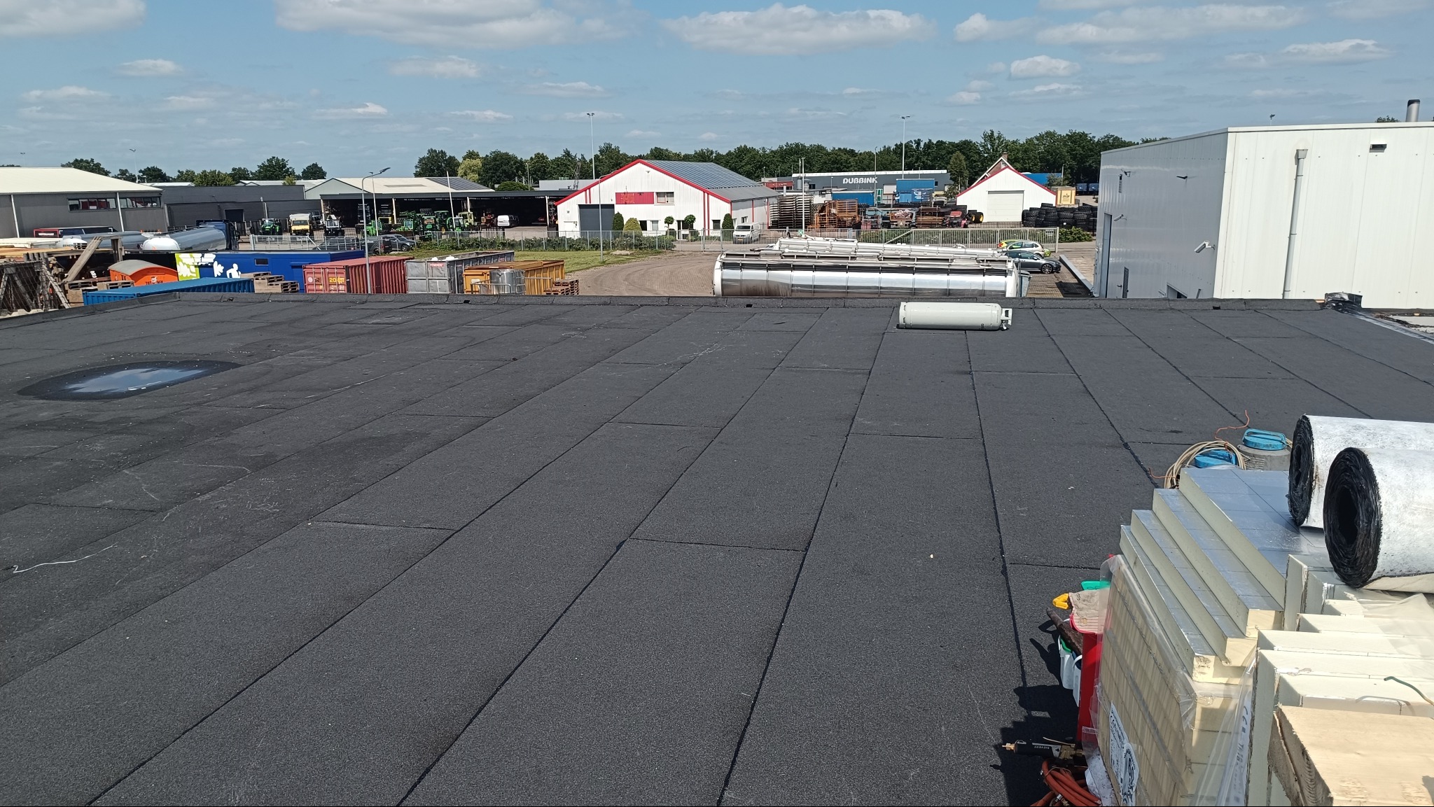 Czarny, płaski dach pokryty papą termozgrzewalną w trakcie prac. Widoczne materiały izolacyjne i zgrzewarka. W tle zabudowania gospodarcze i błękitne niebo.