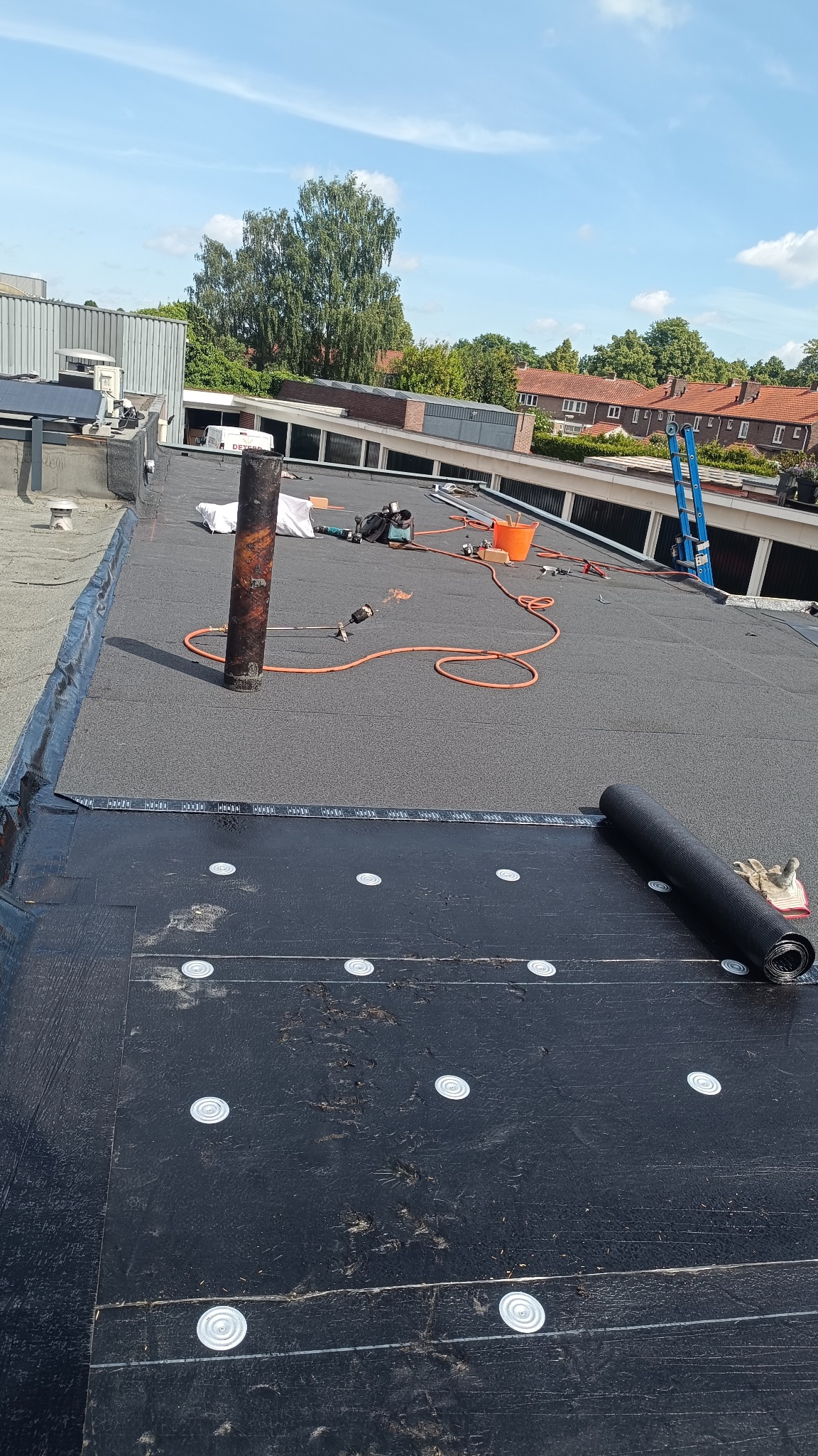 Płaski dach pokryty papą termozgrzewalną z widocznymi narzędziami i niebieską drabiną w tle. Prace dekarskie w toku, widoczny komin i rolka papy.