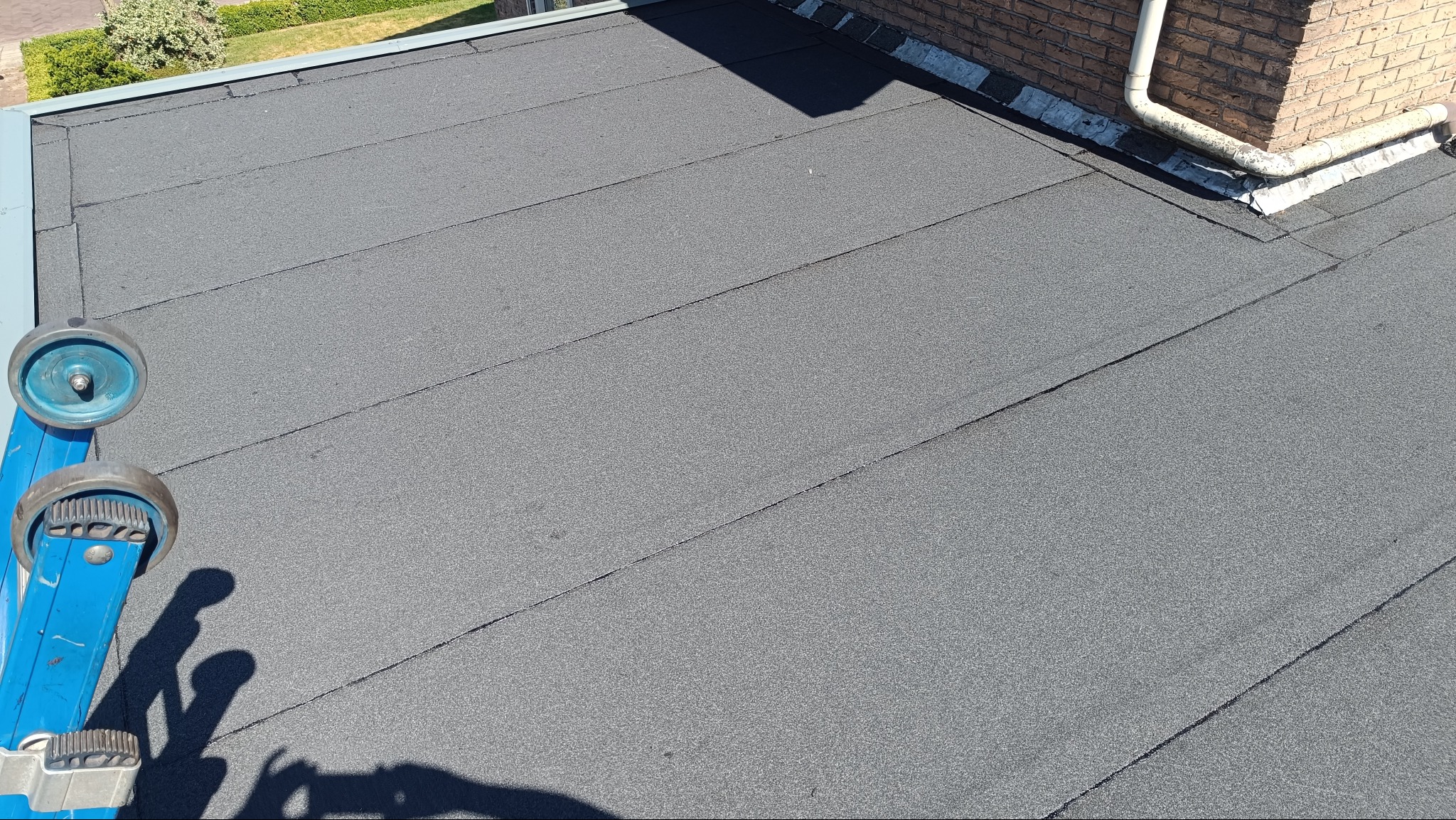 Szary dach pokryty papą z widocznym fragmentem drabiny dekarskiej z rolkami, fragmentem ściany budynku i rynną. Układ połaci dachowych z wyraźnymi liniami łączeń.