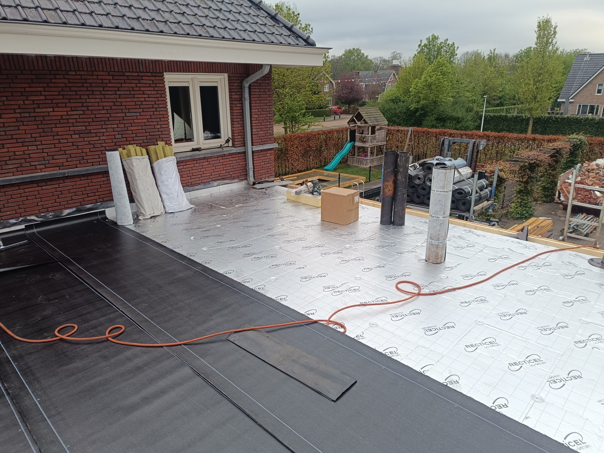 Dach w trakcie krycia papą termozgrzewalną na warstwie izolacyjnej Recticel. Widoczne rolki papy i wełny mineralnej, narzędzia i fragment domu z cegły.