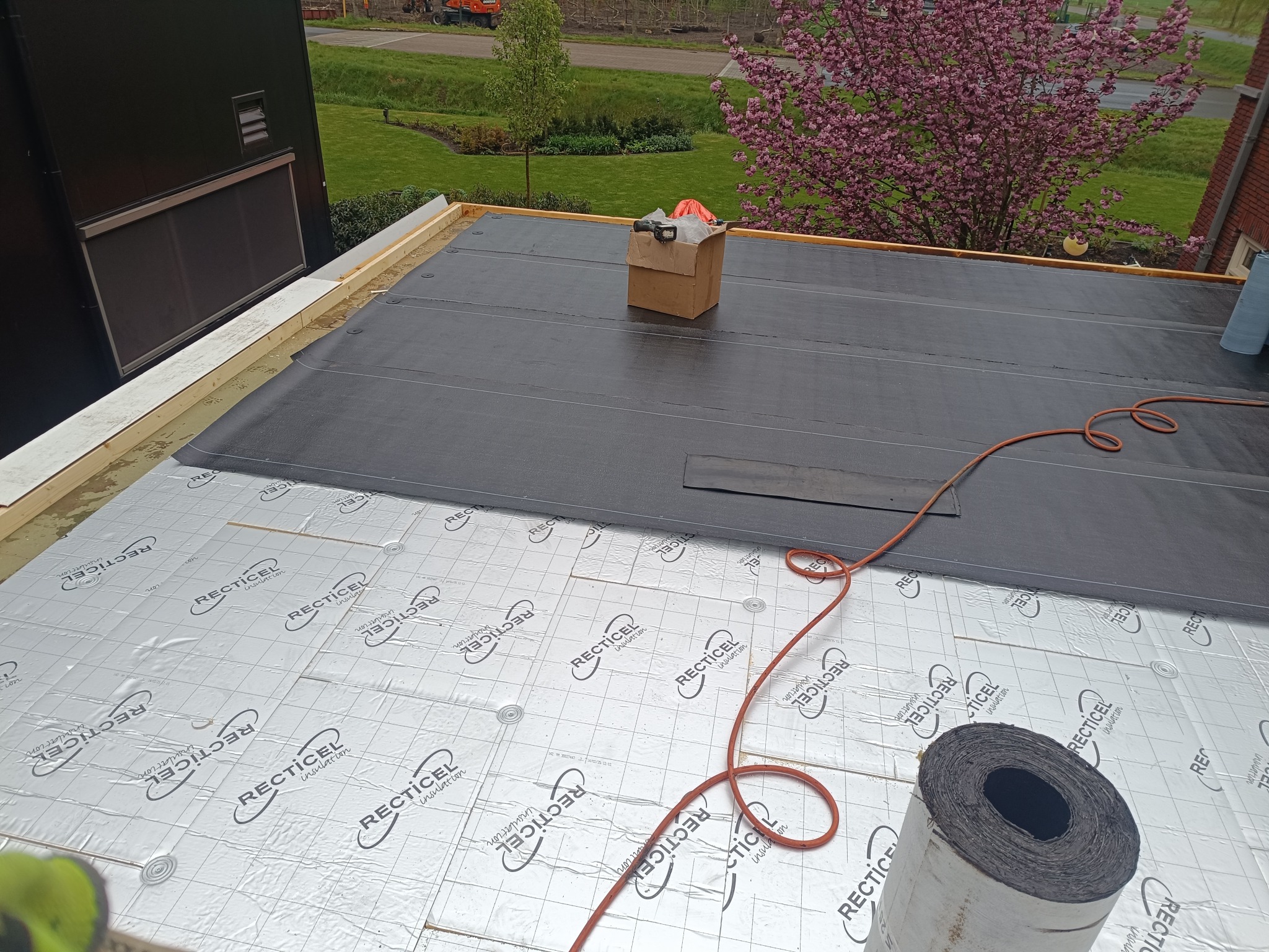 Montaż izolacji podłoża dachu z folią aluminiową Recticel, częściowo pokrytej czarną membraną. Widoczne narzędzia i rolka papy. W tle zieleń drzew i budynek.