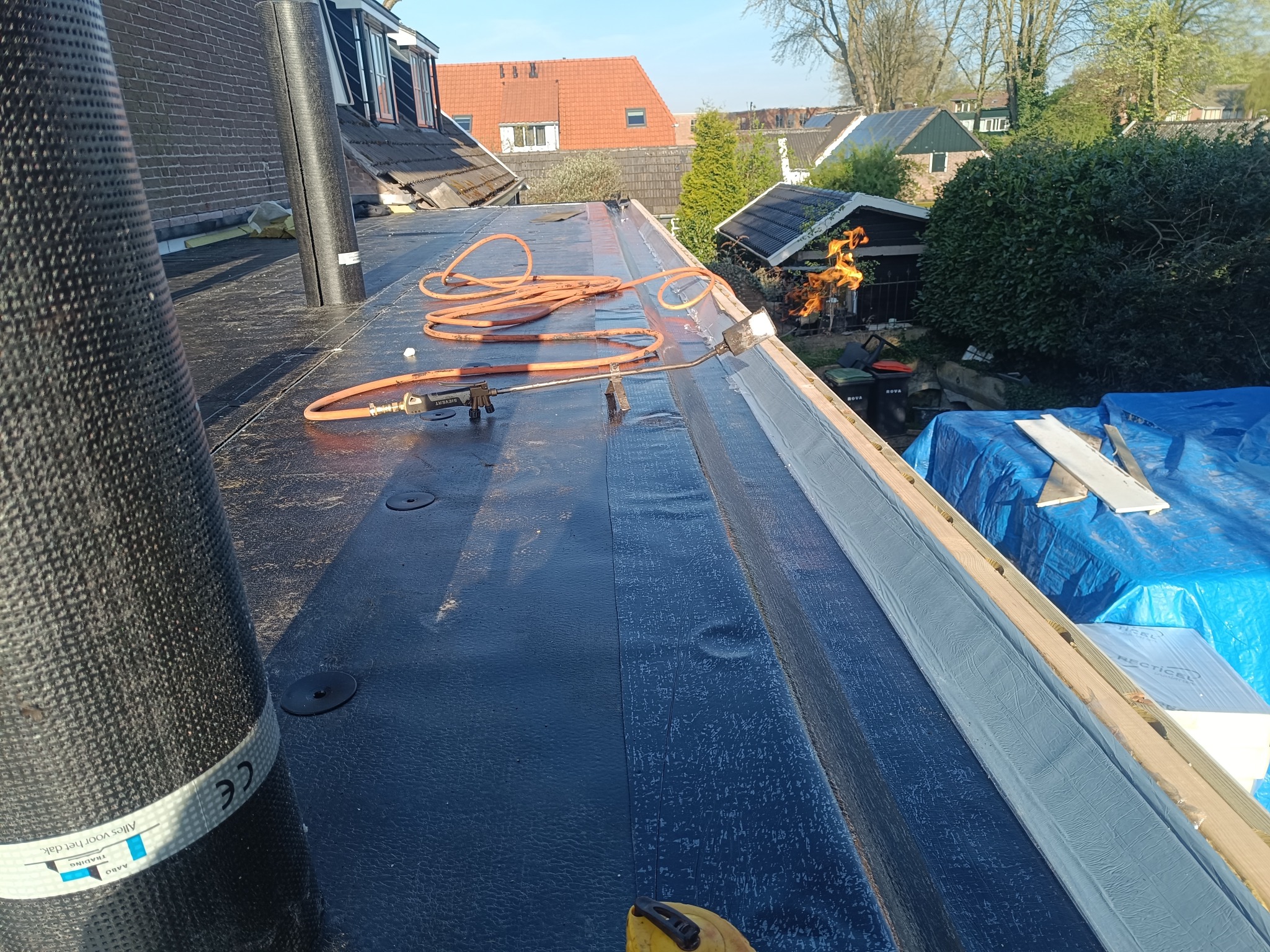 Prace dekarskie na płaskim dachu: papa termozgrzewalna układana palnikiem. W tle zabudowa jednorodzinna i ogrodowa architektura.