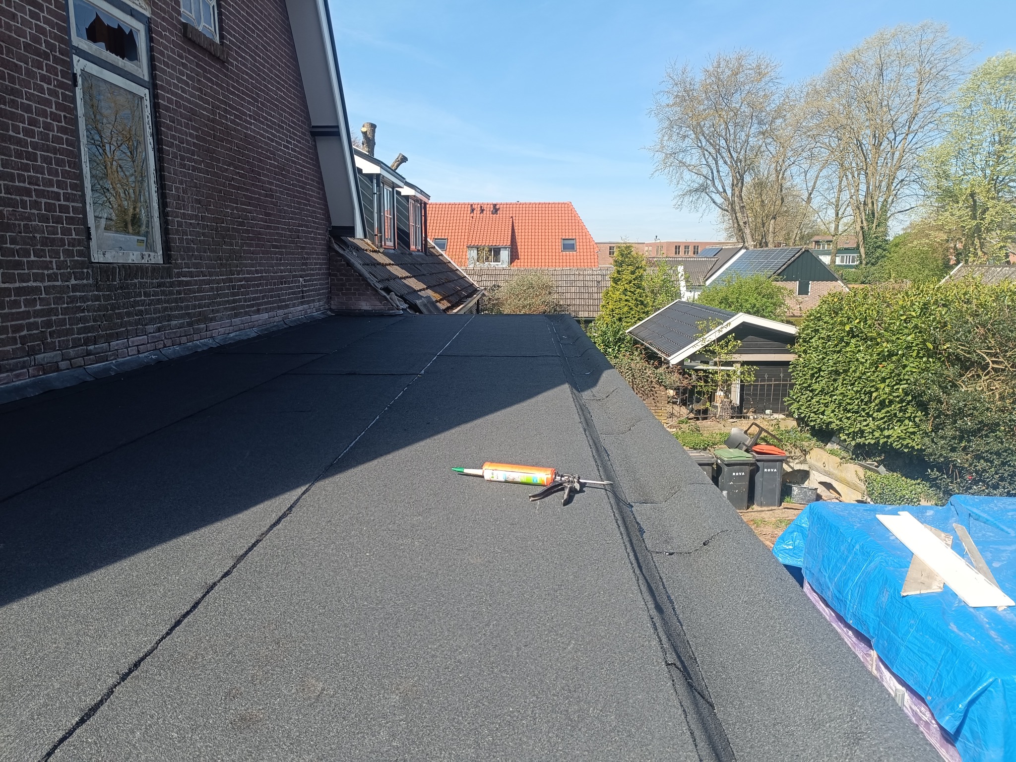 Czarny, płaski dach z papy w słoneczny dzień. Widoczny aplikator do uszczelnień. W tle budynki mieszkalne i zieleń. Uszkodzone okno na ścianie budynku.