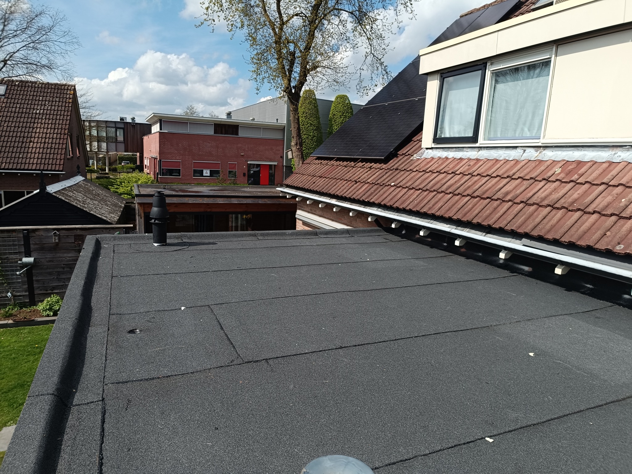 Czarny, płaski dach pokryty papą na tle budynków i zieleni. Widoczne łączenia papy, komin wentylacyjny i fragment dachu z dachówki ceramicznej.