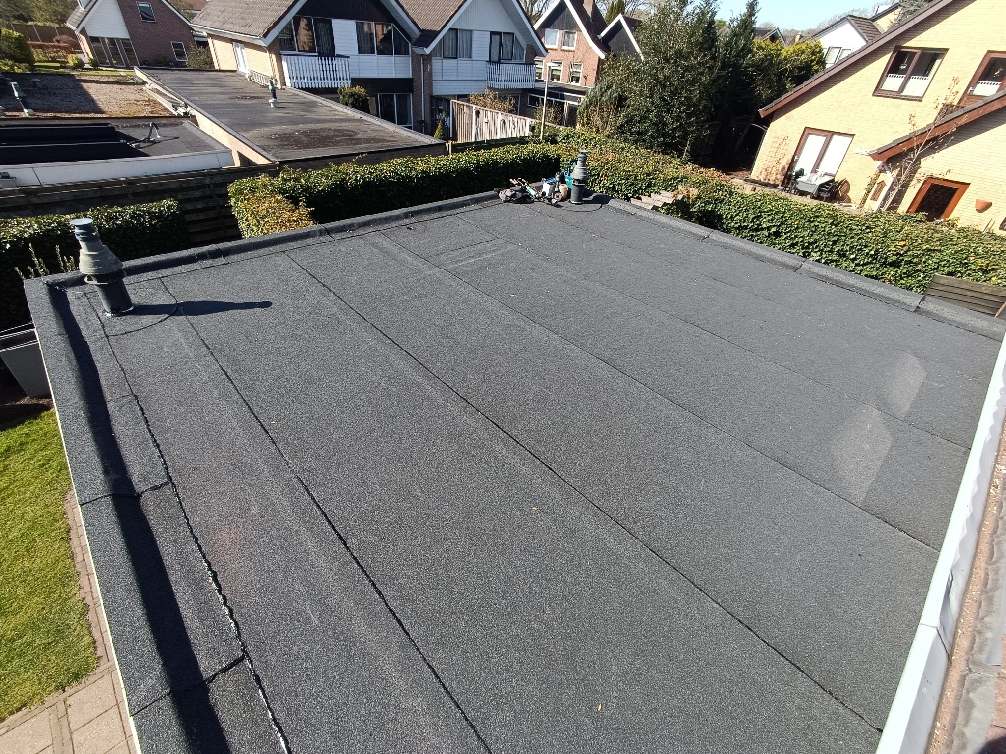 Widok z góry na płaski dach pokryty papą, z widocznymi kominami i narzędziami dekarskimi. W tle zabudowa jednorodzinna i zieleń.